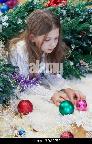 Enfant sur un tapis de fourrure soulevant des ornements verts et roses au milieu de boules et décorations de Noël éparpillées. Banque D'Images