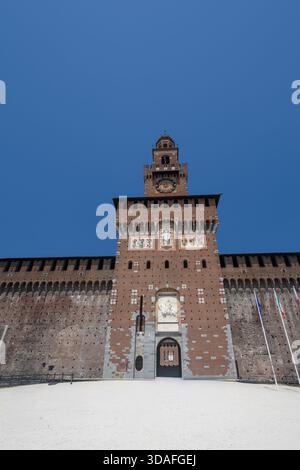 Impressionnante façade médiévale de la forteresse avec une tour de l'horloge centrale, des mosaïques décoratives et des reliefs en pierre. Grande cour ensoleillée et cadre de mâts de drapeau Banque D'Images