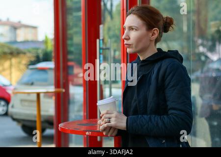 Une jeune femme décontractée aux cheveux roux tient une tasse à emporter, se tient pensivement à l'arrêt de bus rouge avec les bras croisés dans un cadre urbain de jour Banque D'Images