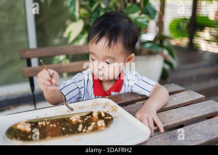 Garçon tout-petit asiatique utilisant la cuillère pour manger le dessert sur le plateau tout en étant assis à la table en bois dans l'environnement de café de jardin Banque D'Images