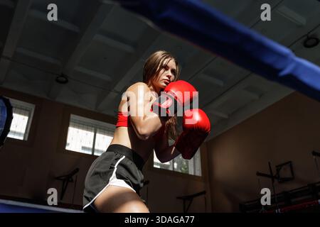 Jeune femme boxeur dans des gants rouges de formation dans l'anneau de gymnastique avec une position de garde ciblée montrant la discipline de force et la compétition Banque D'Images