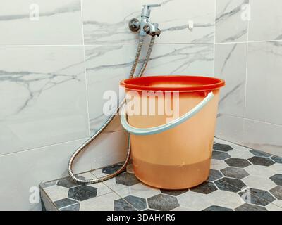 Seau en plastique sous robinet chromé moderne et pomme de douche dans la salle de bains carrelée avec marbre et sol hexagonal Banque D'Images