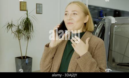 Femme élégante parlant sur son smartphone dans un showroom de voiture à côté d'un véhicule Banque D'Images
