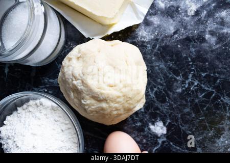 Pâte fraîche maison placée au centre d'un comptoir de cuisine sombre, entourée d'ingrédients de cuisson, y compris des œufs, de la farine, du beurre et du sucre. Flo Banque D'Images