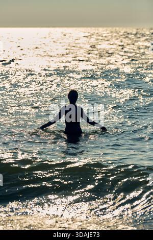Figurine solitaire se dresse jusqu'à la taille dans les eaux scintillantes de la mer Baltique, illuminée par la lumière dorée du coucher du soleil. De douces vagues ondulent autour de la personne, créant t Banque D'Images