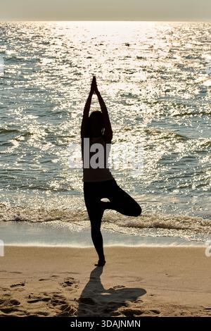 Femme adulte effectue le yoga de pose d'arbre sur la plage de sable avec la toile de fond scintillante de la mer Baltique. La pleine conscience calme et l'équilibre soulignés par des lightti sereins au coucher du soleil Banque D'Images