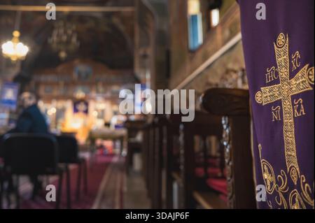Petrosani, Roumanie 22 novembre 2025 : tissu liturgique violet or brodé croix orthodoxe (IHC XC NIKA) intérieur de l'église et bancs flou sur fond Banque D'Images