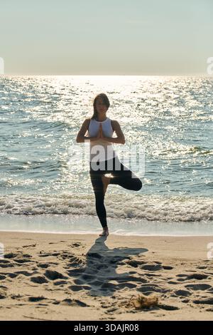 Une femme adulte effectue un exercice de yoga de pose d'arbre sur le rivage de sable de la mer Baltique avec de l'eau calme et la lumière du soleil chatoyante. Scène capture la pleine conscience et tranqui Banque D'Images