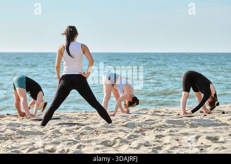 Jeunes femmes pratiquant le yoga de groupe sur la plage de sable au bord de la mer Baltique. Le fond de mer calme et le ciel dégagé améliorent l'activité de fitness en plein air sereine Banque D'Images