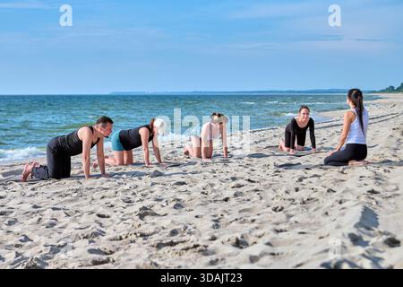 Quatre femmes adultes effectuant des poses de yoga sur la rive sablonneuse de la mer Baltique, guidées par un instructeur. Océan calme et lumière naturelle lumineuse ensemble paisible extérieur ex Banque D'Images