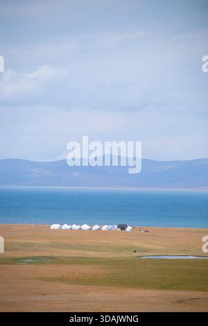 Vue panoramique de yourtes nomades blanches installées sur une plaine herbeuse à côté d'un grand lac bleu avec une chaîne de montagnes en toile de fond sous un ciel nuageux. Banque D'Images