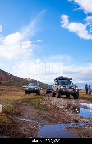 Naryn Region, Kirghizistan - 13 mai 2024 : voitures d'expédition noir et lexus conduisant sur une piste boueuse avec flaque d'eau tandis qu'un groupe de touristes se trouve à proximité Banque D'Images