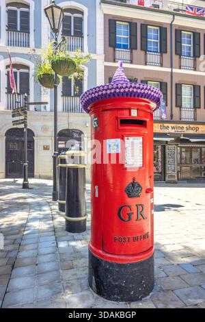 Gibraltar, Royaume-Uni - 10 septembre 2024 : boîte postale victorienne de conception britannique standard. Une boîte aux lettres rouge ornée d'un bonnet tricoté repose sur un pavé Banque D'Images
