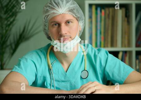 Un médecin masculin ou infirmier en uniforme médical avec masque et bonnet chirurgical est assis dans son bureau. Stéthoscope autour de son cou. Regardez la caméra de façon calme et sérieuse. Concept de diagnostic médical. Banque D'Images