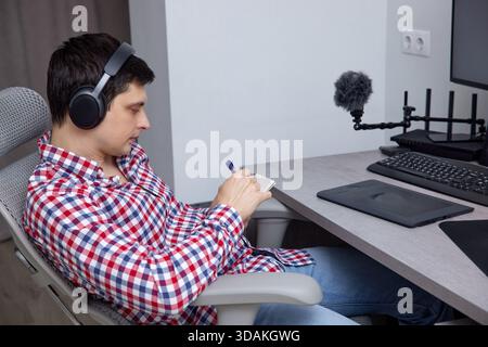 Homme portant des écouteurs assis à un bureau ergonomique, écrivant des notes dans un ordinateur portable Banque D'Images