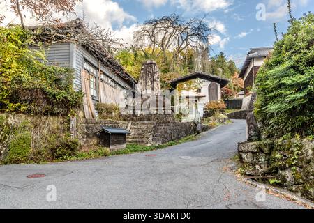 Une pierre commémorative sur un escalier en pierre sur la route d'accès à Tsumago-juku, sur fond de feuillage automnal, avec des bâtiments résidentiels en arrière-plan ; cette photo grand angle capture la transition de la périphérie moderne au centre-ville historique de Nagiso, préfecture de Nagano, Japon Banque D'Images
