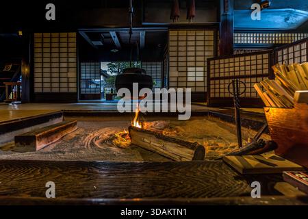 Un foyer irori traditionnel dans une machiya historique à Nagiso, Nagano : une grande bouilloire tetsubin en fonte est suspendue au-dessus du feu ouvert sur une chaîne de fer attachée aux poutres du toit. Pendant la période Edo, l’irori était la principale source de chaleur et la principale zone de cuisson des maisons japonaises, tout en servant de centre social pour la famille. En arrière-plan se trouvent des salles de tatami avec des portes shoji et une vue sur le jardin. Nagiso, Nagano, Japon Banque D'Images