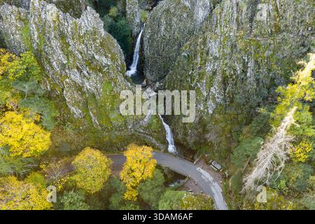 Vue aérienne de cascades plongeant dans des falaises couvertes de mousse, embrassées par des arbres d'automne et une route sinueuse à Pozo do Inferno, Manteigas, po Banque D'Images
