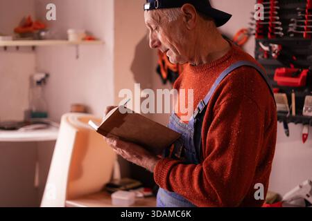 Mécanicien masculin âgé ayant une conversation téléphonique avec le client écrivant des commandes dans le bloc-notes à l'atelier. vue latérale Banque D'Images