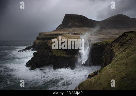 Vue sur la puissante cascade Mulafossur cascades dans la mer turbulente depuis des falaises escarpées sous un ciel spectaculaire, Sorvagur, Vagar, îles Féroé. Banque D'Images