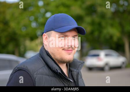 Portrait en plein air d'un jeune homme confiant souriant tout en portant une casquette bleue de baseball et un gilet noir. Banque D'Images