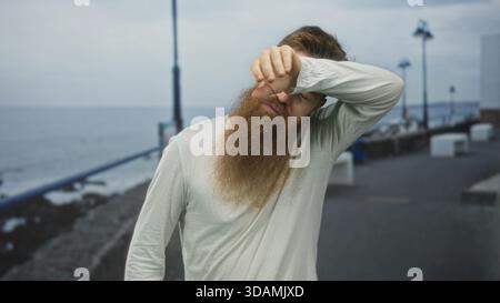 Homme barbu en chemise blanche essuie la sueur du front sur la promenade chaude et ensoleillée de bord de mer ; soulagement. Banque D'Images