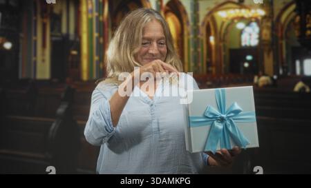 Femme senior blonde pointe le doigt à la boîte cadeau emballée avec la main visible tout en souriant et tenant présent à l'intérieur du bâtiment orné ; contentement chaleureux. Banque D'Images