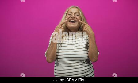 Femme senior pointant les doigts sur les joues et souriant avec les dents dans un portrait de studio sur un fond magenta ; joie ludique. Banque D'Images