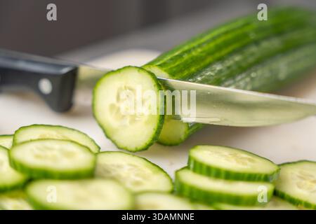 Un couteau de cuisine tranchant coupe un concombre sur une planche. Les tranches fraîches tombent vers l'avant, capturant le moment de la préparation des aliments et des légumes croquants fres Banque D'Images
