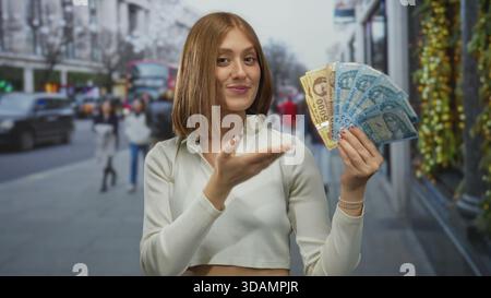 Jeune femme blonde tenant des billets de banque hongrois en forint souriant dans une rue urbaine avec un paysage urbain en arrière-plan, mettant en valeur la monnaie à l'extérieur. Banque D'Images
