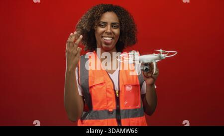 Femme tenant un drone et faisant une pompe de poing avec la main visible tout en souriant dans le studio avec fond rouge ; accomplissement d'excitation. Banque D'Images