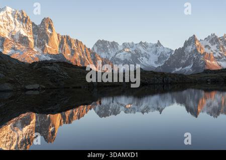 Vue sur les montagnes dentelées et enneigées reflétant le lac serein, peint avec les teintes chaudes du soleil couchant, Lacs des Cheserys, Auvergne-Rhône-A. Banque D'Images