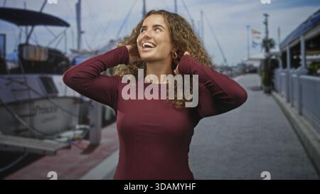 Femme souriant et plaçant les mains derrière la tête tout en ajustant les cheveux bouclés à côté des yachts amarrés sur un quai de marina, regardant vers le haut ; joie insouciante. Banque D'Images