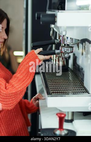 Concentrez-vous sur une femme vêtue d'une tenue orange qui fixe la poignée à la machine à espresso avec une inviolabilité visible. Banque D'Images