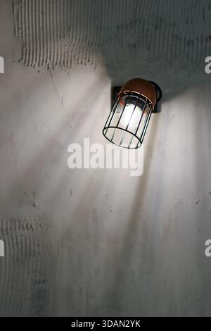 Vue détaillée du luminaire industriel monté sur le mur, abat-jour brun, cadre noir, ampoule en cage émettant de la lumière. Banque D'Images