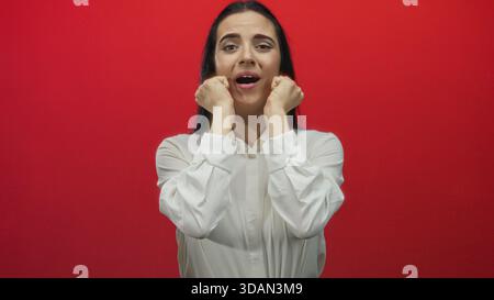 Femme exprimant l'excitation avec ses mains près du visage contre un mur rouge vif, véhiculant l'émotion intense et l'énergie, debout dans une chemise blanche, captur Banque D'Images