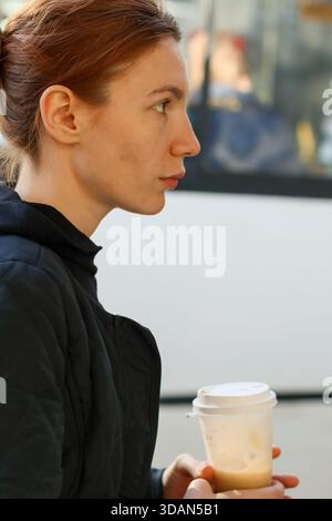 Gros plan d'une jeune femme réfléchie avec queue de cheval serrant le café à emporter près d'un bus public en milieu urbain Banque D'Images