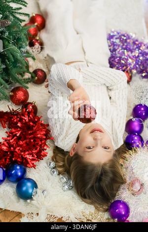 Un enfant en tricot attire l'ornement scintillant plus près tout en s'inclinant sur un tapis doux entouré d'éléments de vacances Banque D'Images