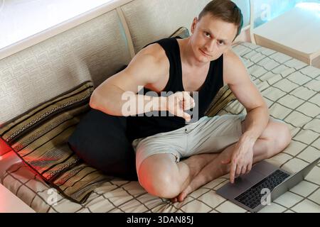 Jeune homme décontracté assis sur le lit, pointant vers le bas et les pouces vers le bas à l'écran de l'ordinateur portable avec un regard désapprobateur Banque D'Images