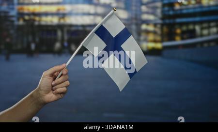 Main tenant le drapeau de la finlande dans la scène de rue de la ville avec fond urbain flou pendant le crépuscule représentant la fierté nationale et la culture. Banque D'Images