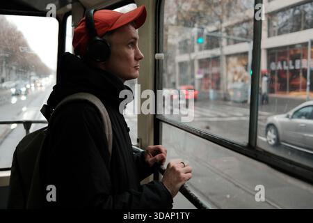 Un homme se tient dans un tramway portant des écouteurs et regarde par la fenêtre la rue pluvieuse. Le moment transmet le concept de trajet urbain et d'humeur de voyage. Banque D'Images