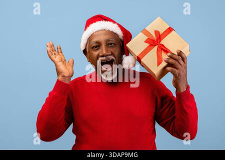 Homme plus âgé en chandail rouge et chapeau de Père Noël tient cadeau tout en célébrant la saison des fêtes Banque D'Images