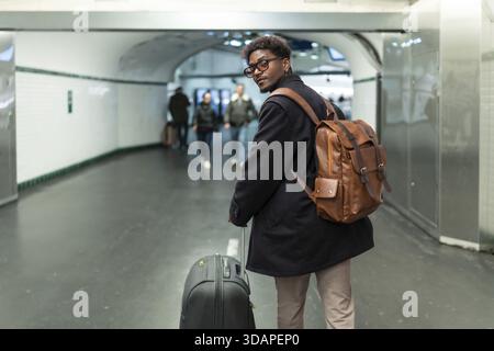 Jeune homme avec un sac à dos en cuir élégant et des bagages à roulettes fait une pause pour regarder en arrière dans une station de métro moderne ou un passage souterrain, se préparant pour Banque D'Images