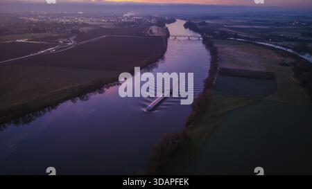 Un cargo se déplace sur le large Danube pendant le coucher du soleil. Le paysage montre des champs et des arbres le long du rivage. Il est le soir et la ville est visible en th Banque D'Images