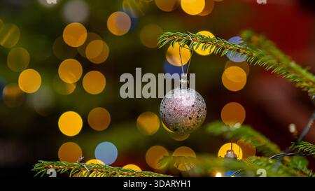 Une boule de Noël argentée pailletée est suspendue à une branche de sapin verte, entourée de lumières bokeh dorées chaudes et colorées pour créer un atmo festif Banque D'Images