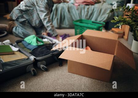 Jeune femme agenouillée sur la valise d'emballage de plancher avec des vêtements et des objets personnels, boîte en carton ouverte et panier à linge à proximité, se préparant pour le voyage ou le déménagement Banque D'Images