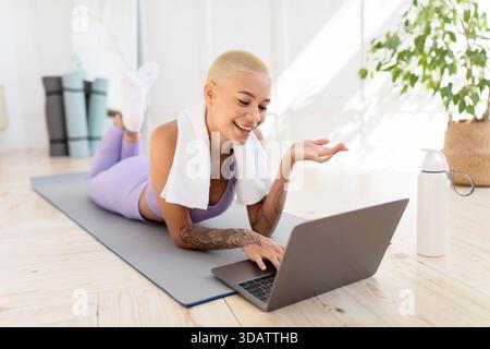 Heureuse dame dans la tenue de sport couchée sur le tapis de fitness, parler avec l'entraîneur en ligne sur ordinateur portable à la maison Banque D'Images
