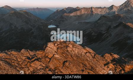 Vue sur une figure solitaire debout au sommet de pics déchiquetés et ensoleillés surplombant des lacs tranquilles et réfléchissants nichés au milieu des vastes montagnes escarpées de Kloste Banque D'Images