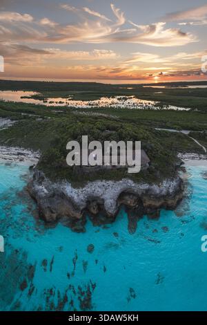Vue aérienne d'un affleurement rocheux surmonté d'arbres denses et d'un bâtiment, bordé de sable blanc et d'eaux turquoises au coucher du soleil, Eleuthera, Bahamas. Banque D'Images