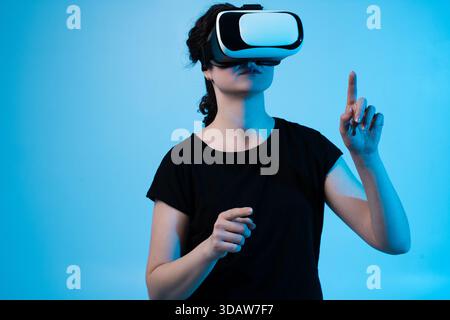 Femme portant casque VR interagissant avec l'environnement virtuel et pointant vers l'avant sur fond bleu Banque D'Images